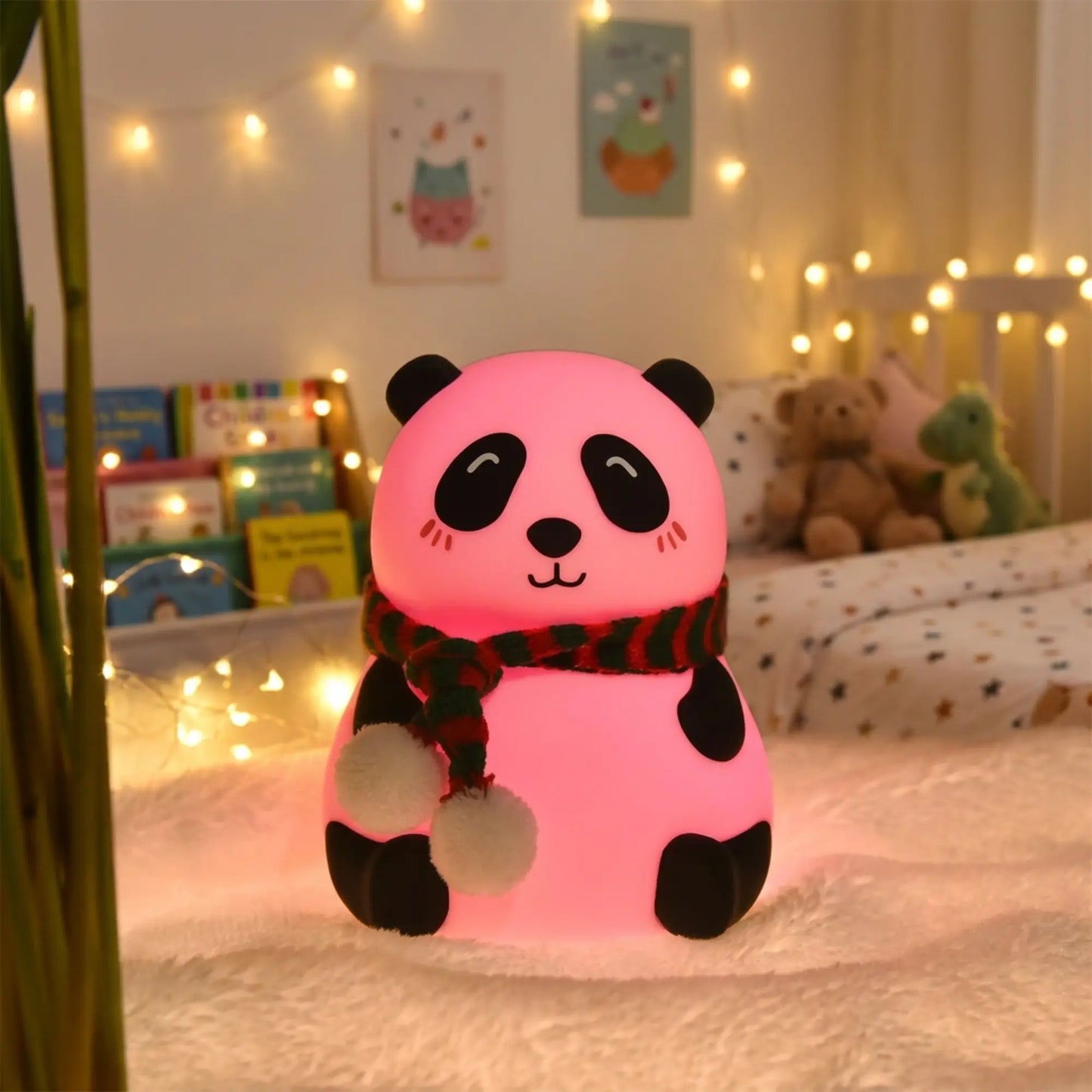 A pink panda-shaped night light glowing with a warm glow, wearing a red and black striped scarf with white pom-poms, set on a white fluffy rug in a cozy, warmly lit children's bedroom.
