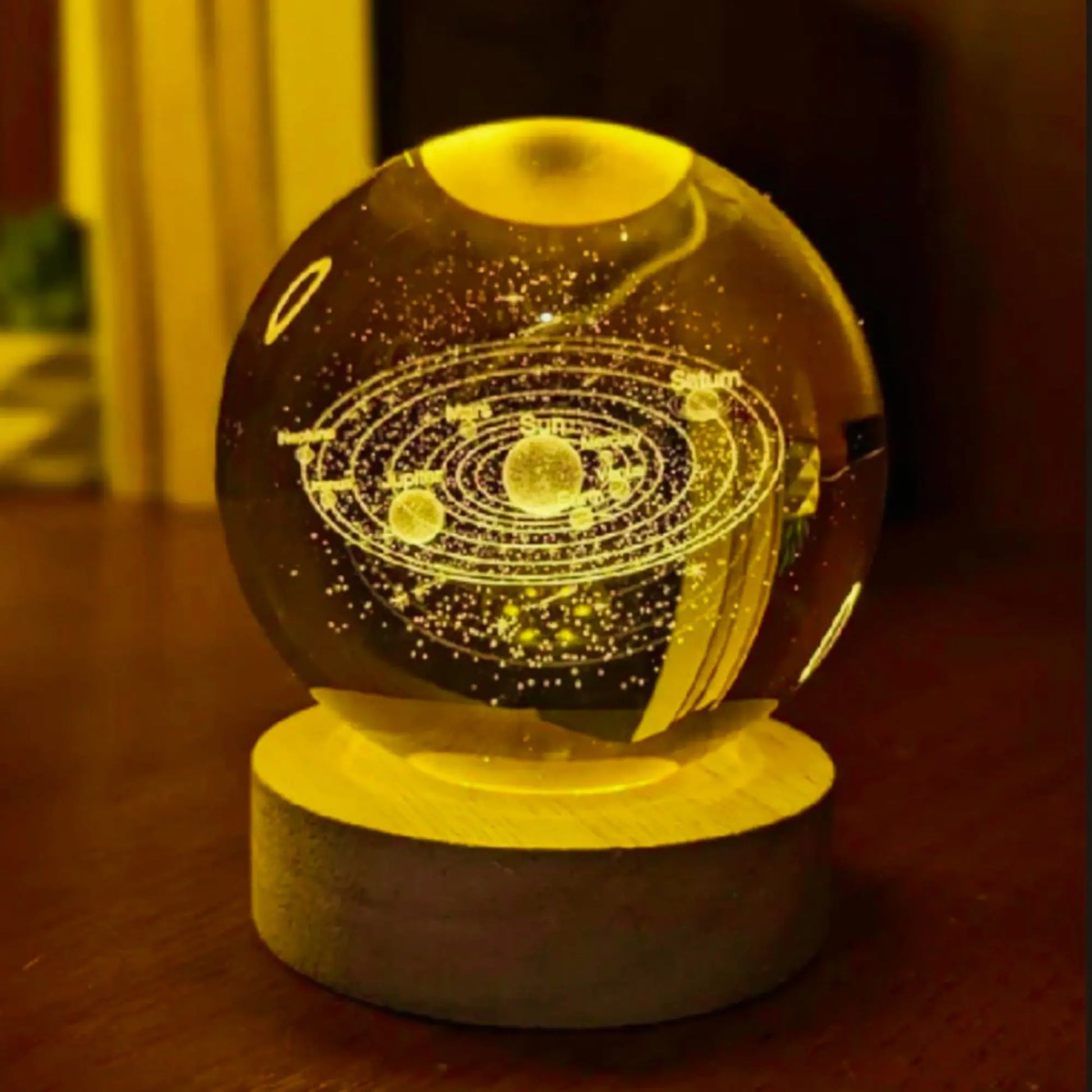 A glowing crystal ball night light with an etched solar system design, resting on a round wooden base on a dark wooden table.