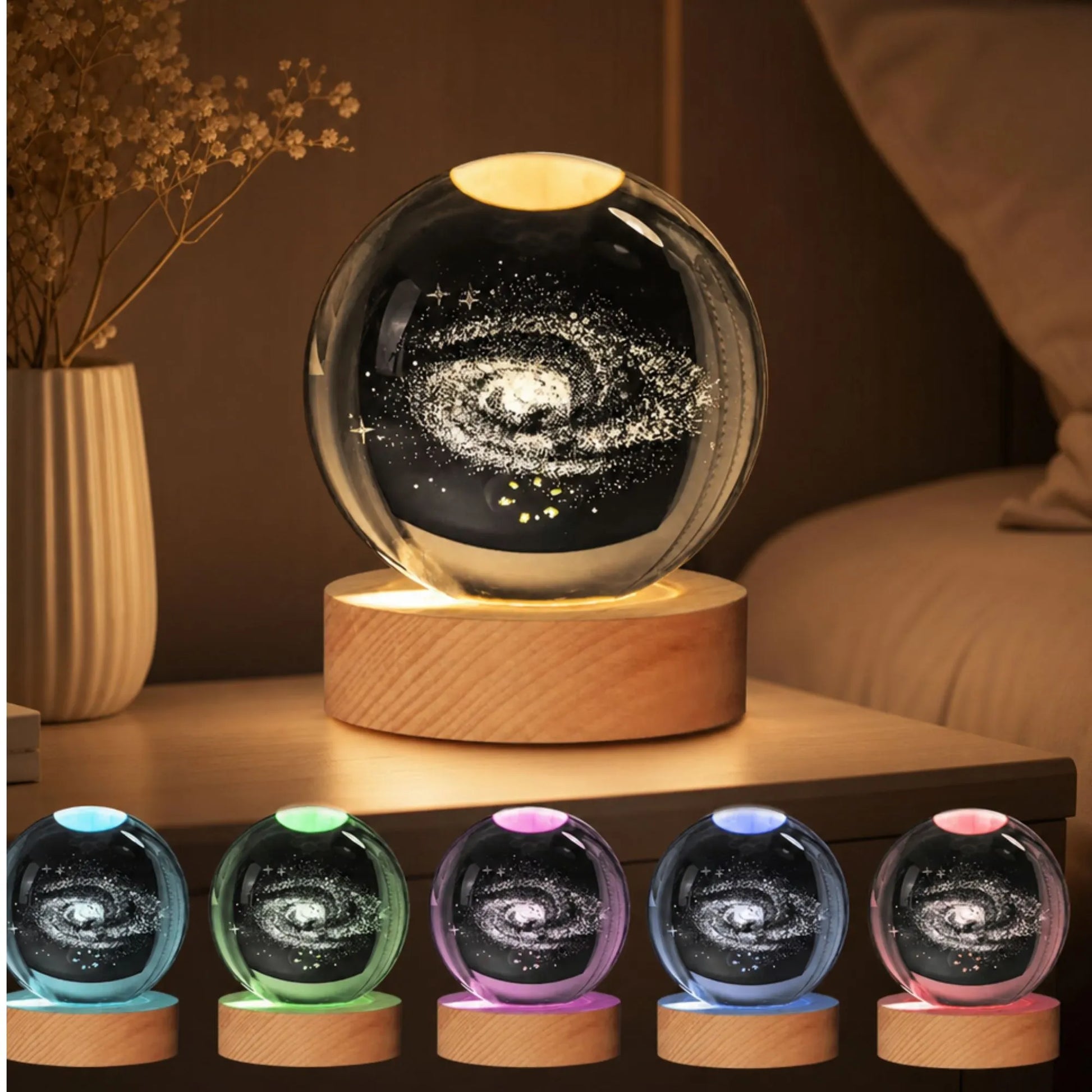 A glowing spherical galaxy night light on a wooden base, shown lit on a bedside table with a bed and vase in the background. Five smaller color variants are lined up along the bottom.