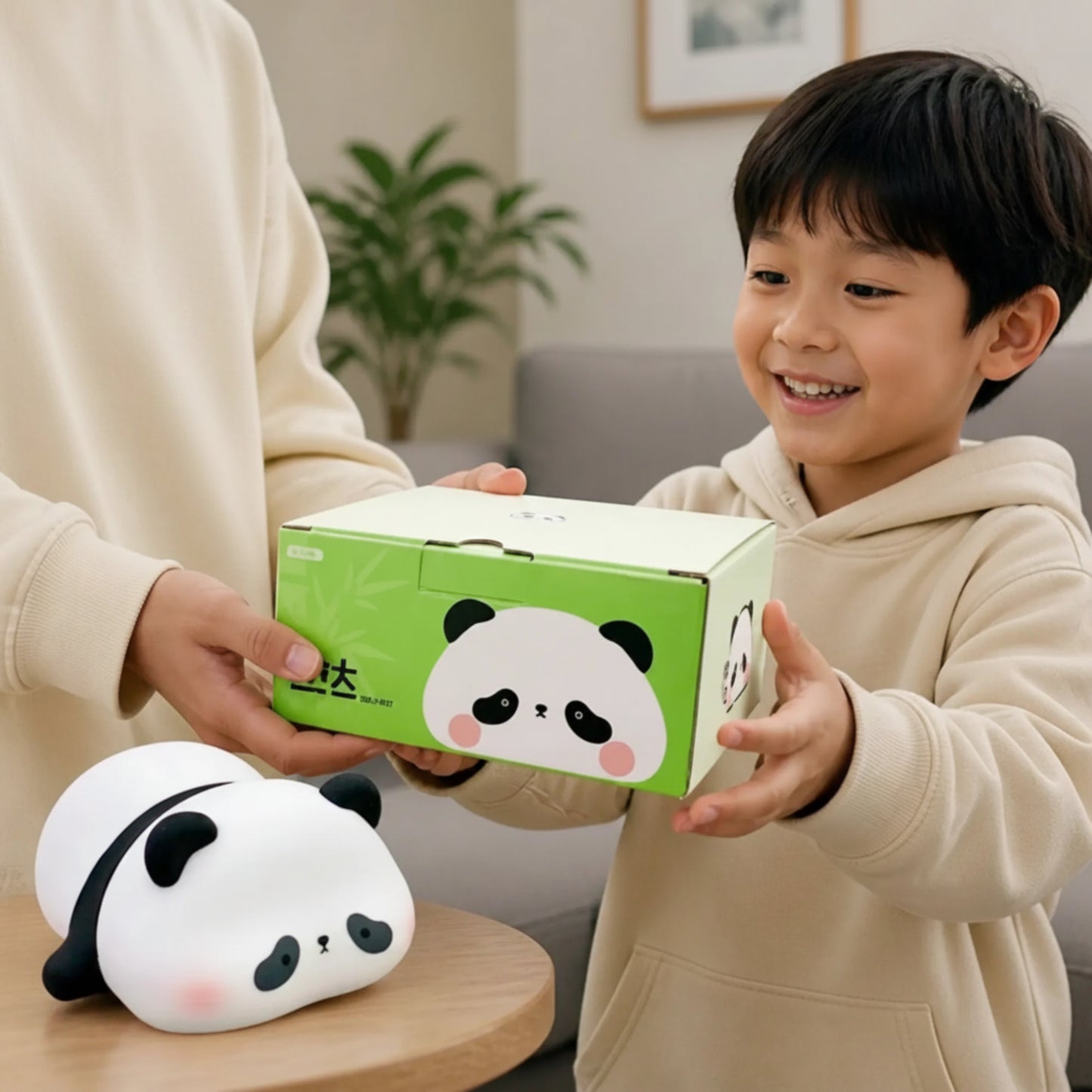 A smiling young boy in a beige hoodie holds a green and white panda-themed product box while an adult in a matching beige sweatshirt assists. A white panda plush toy rests on a small wooden table in the foreground, set against a softly blurred living room background.