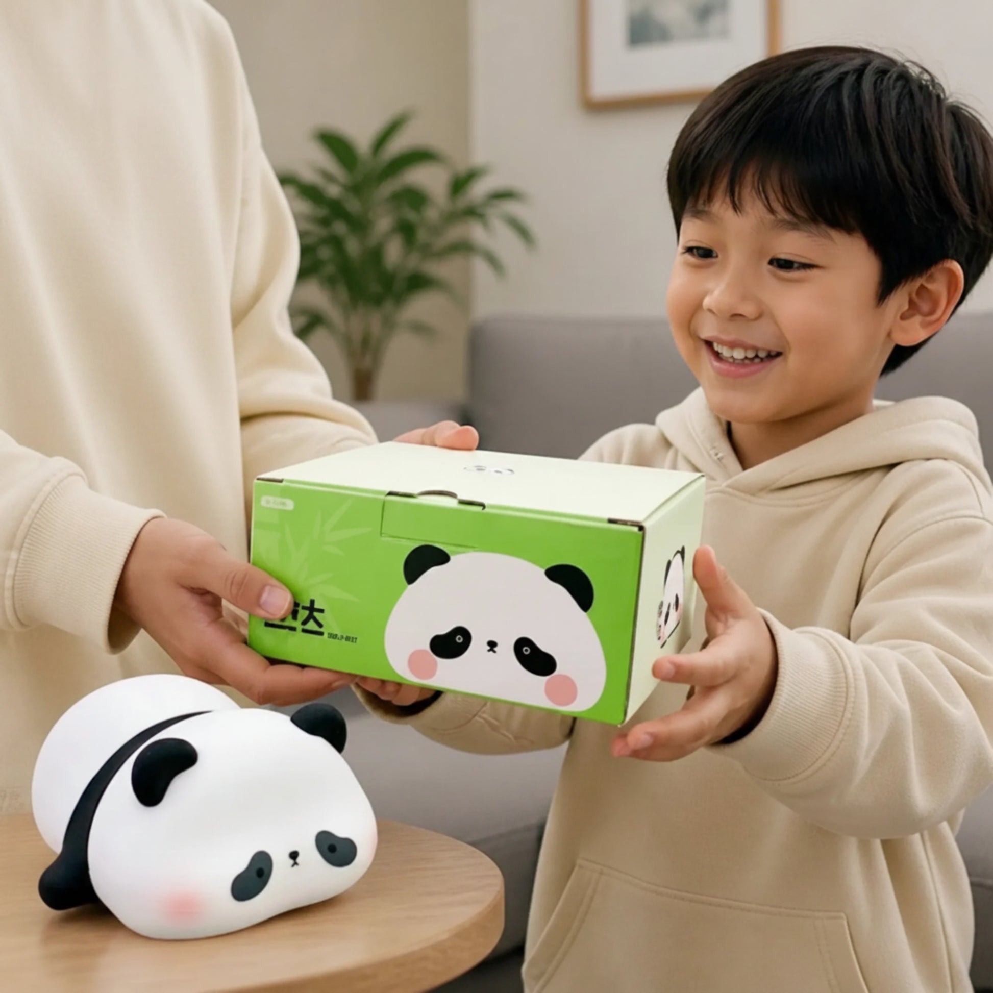 A smiling young boy in a beige hoodie holds a green and white panda-themed product box while an adult in a matching beige sweatshirt assists. A white panda plush toy rests on a small wooden table in the foreground, set against a softly blurred living room background.